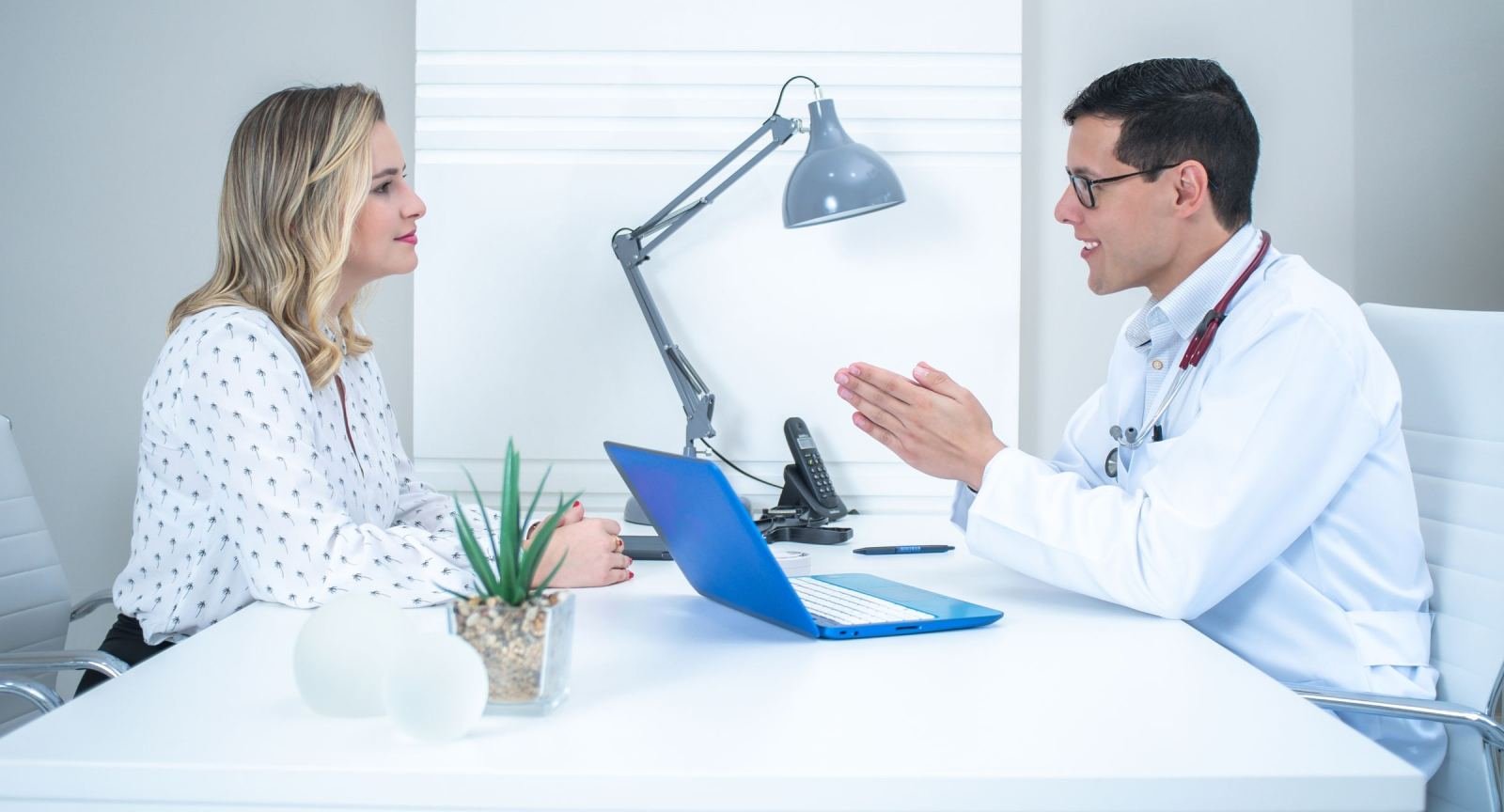 A woman and a clinician in a white coat talk across a desk with a laptop