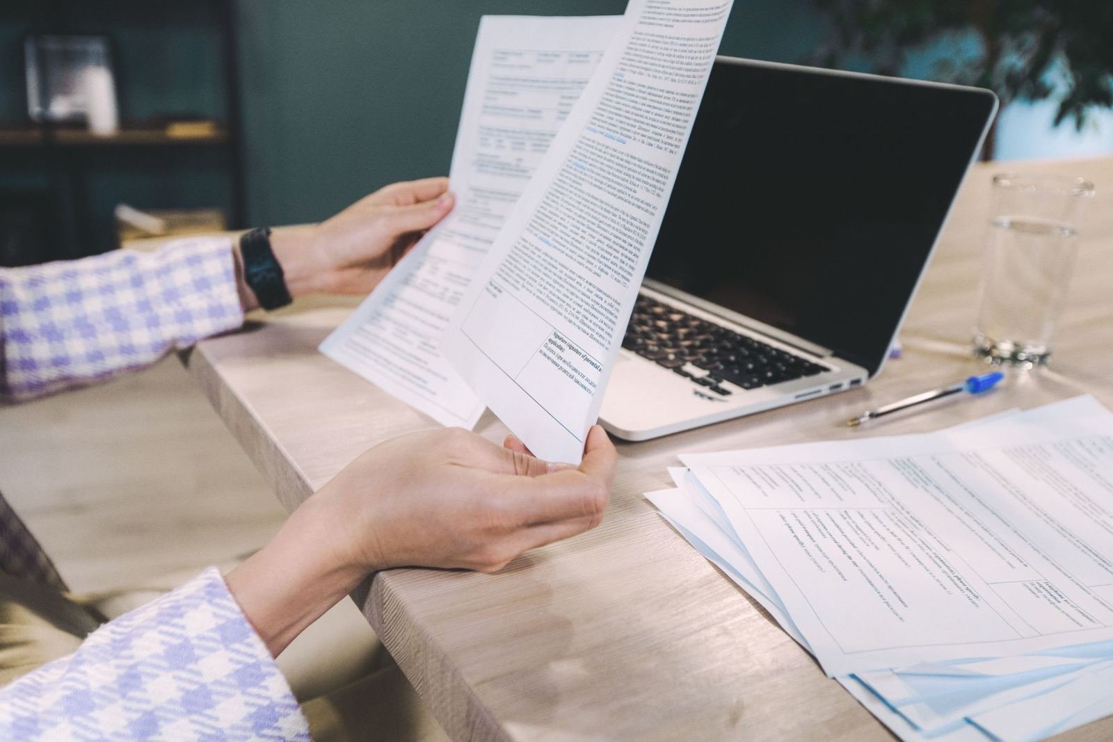 Hands holding and reviewing printed documents next to a laptop and glass of water