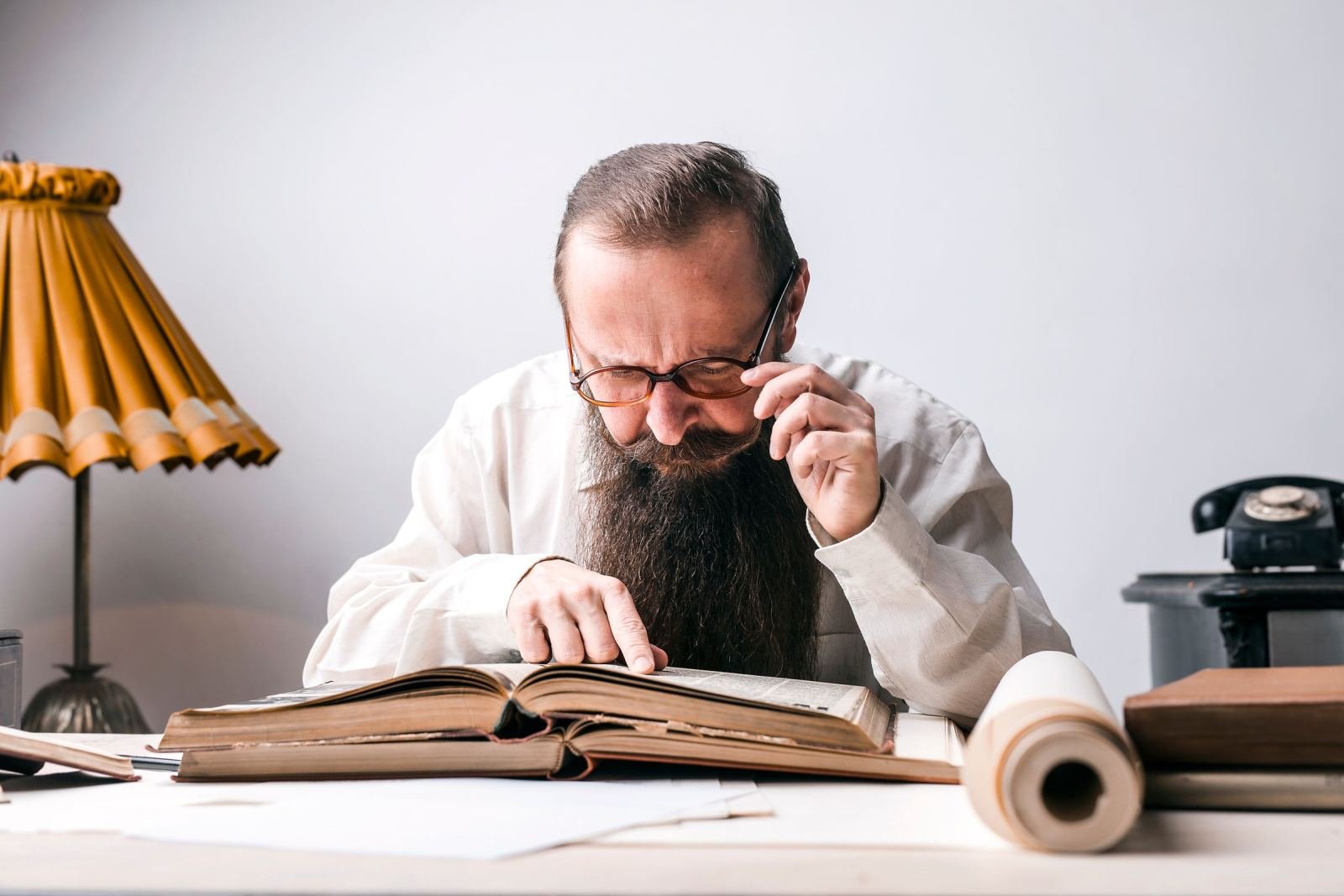 A man with a beard and glasses reads a large open book at a desk