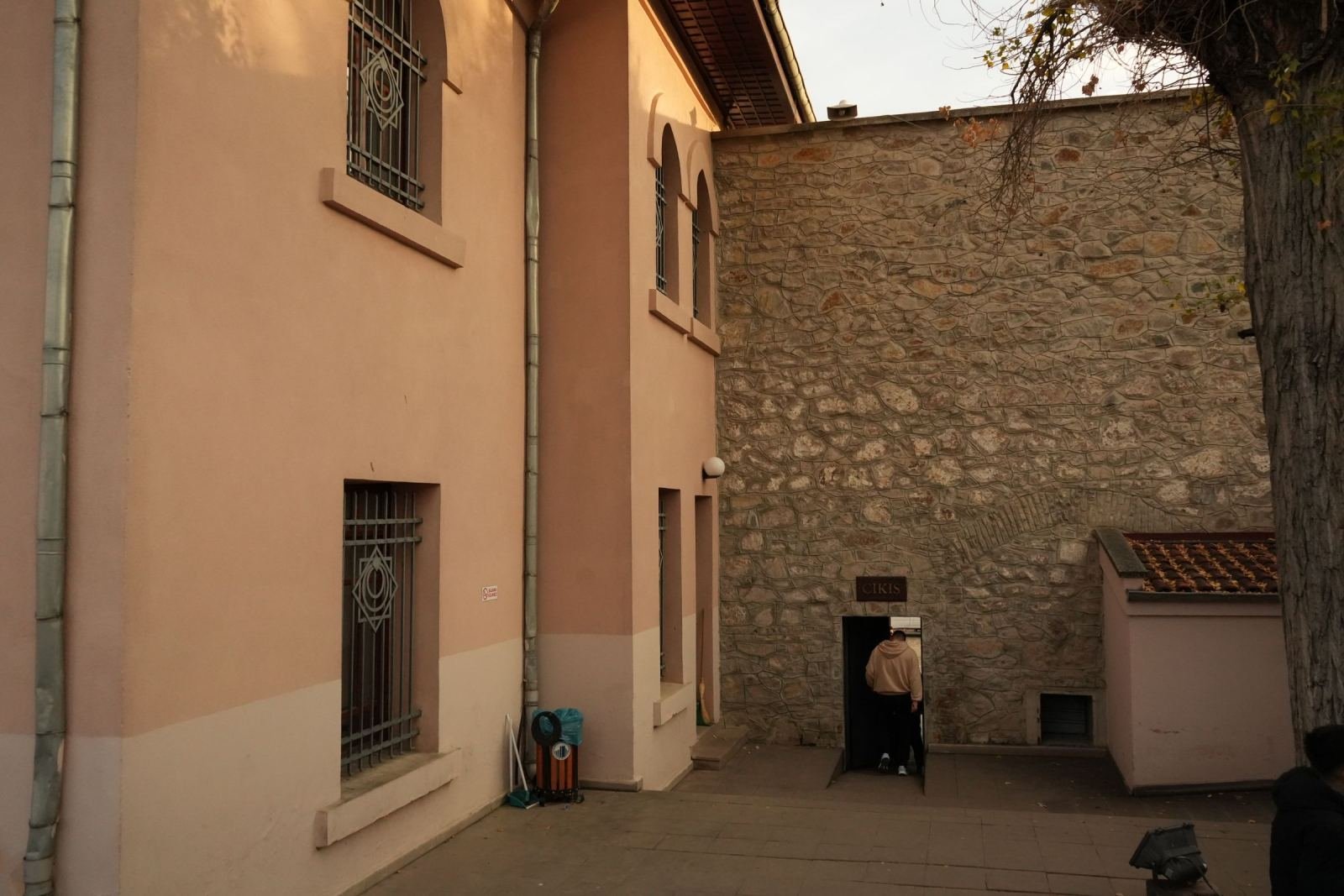 A woman steps through a door into a stone house beside a pink wall.