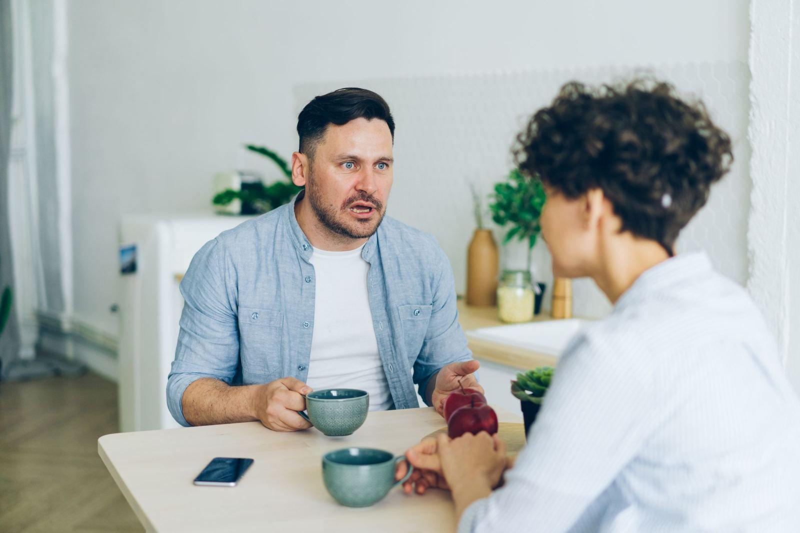 Two people, a man and a woman, sit at a table. They talk while holding mugs of coffee.