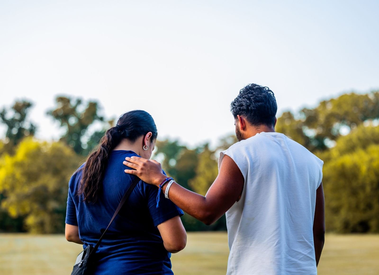One person puts a hand on the other's shoulder as they walk side by side in a park.