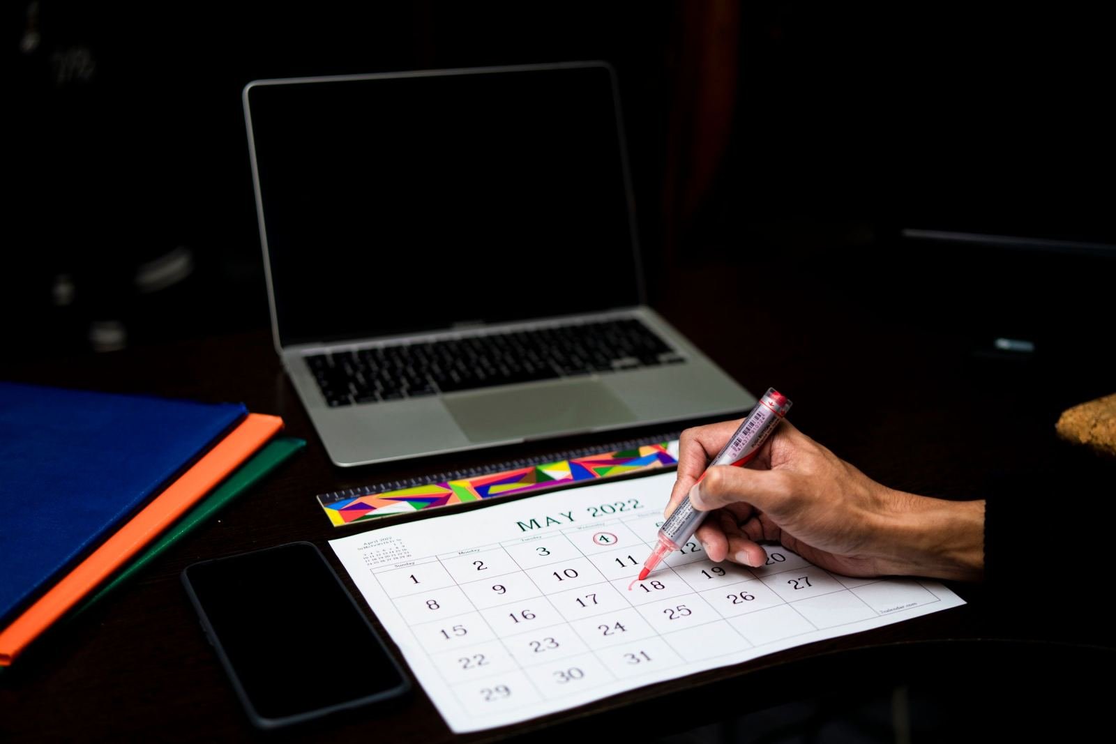 A hand circles a date on a May 2022 calendar next to a laptop and smartphone