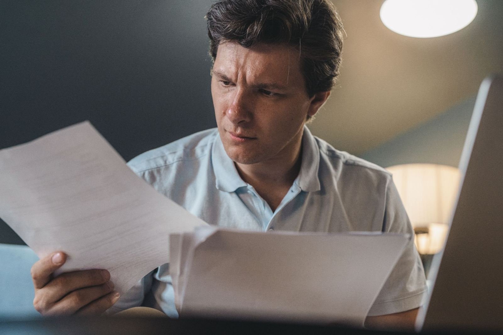 A man with a worried look reads a paper near his laptop