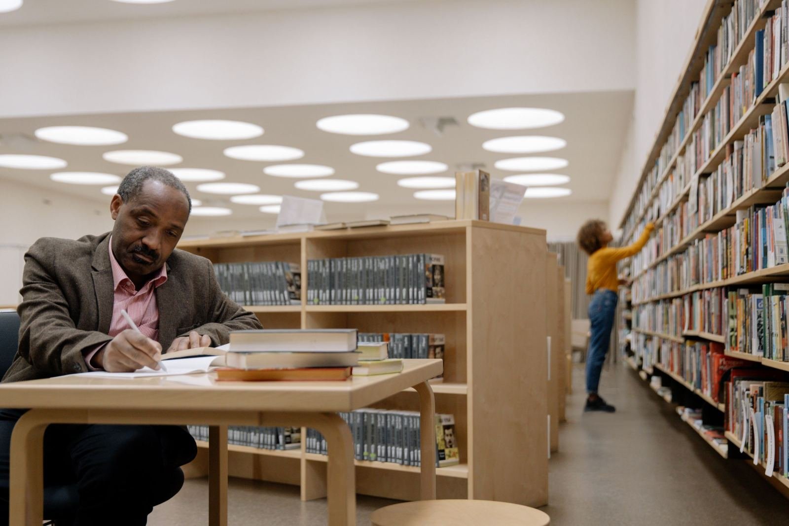 A man writes in a notebook at a library table with books stacked nearby