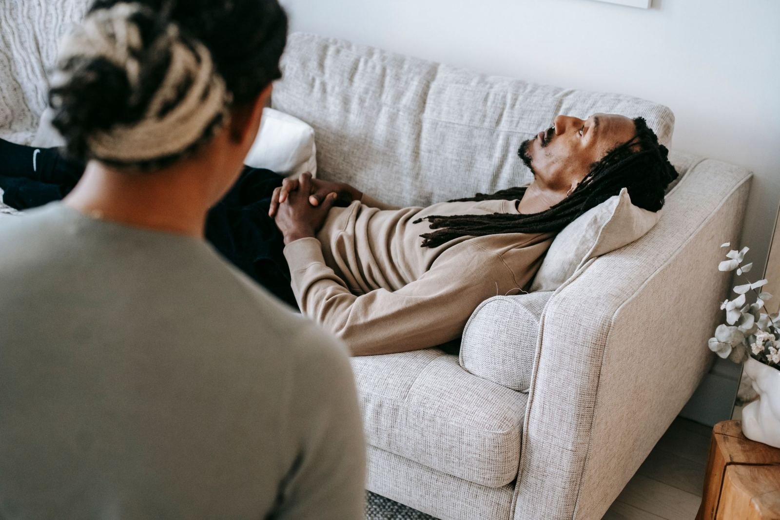 A man with dreadlocks lies on a sofa while talking to a person seen from behind