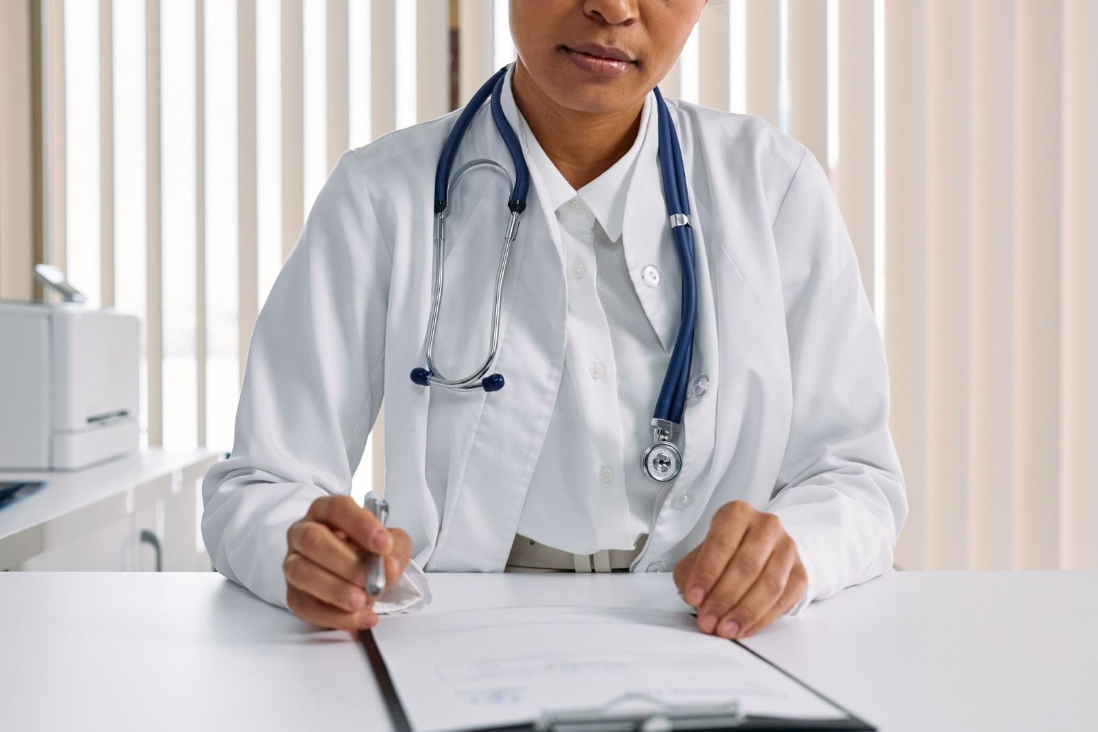 A doctor in a white coat and stethoscope sits at a desk with a clipboard