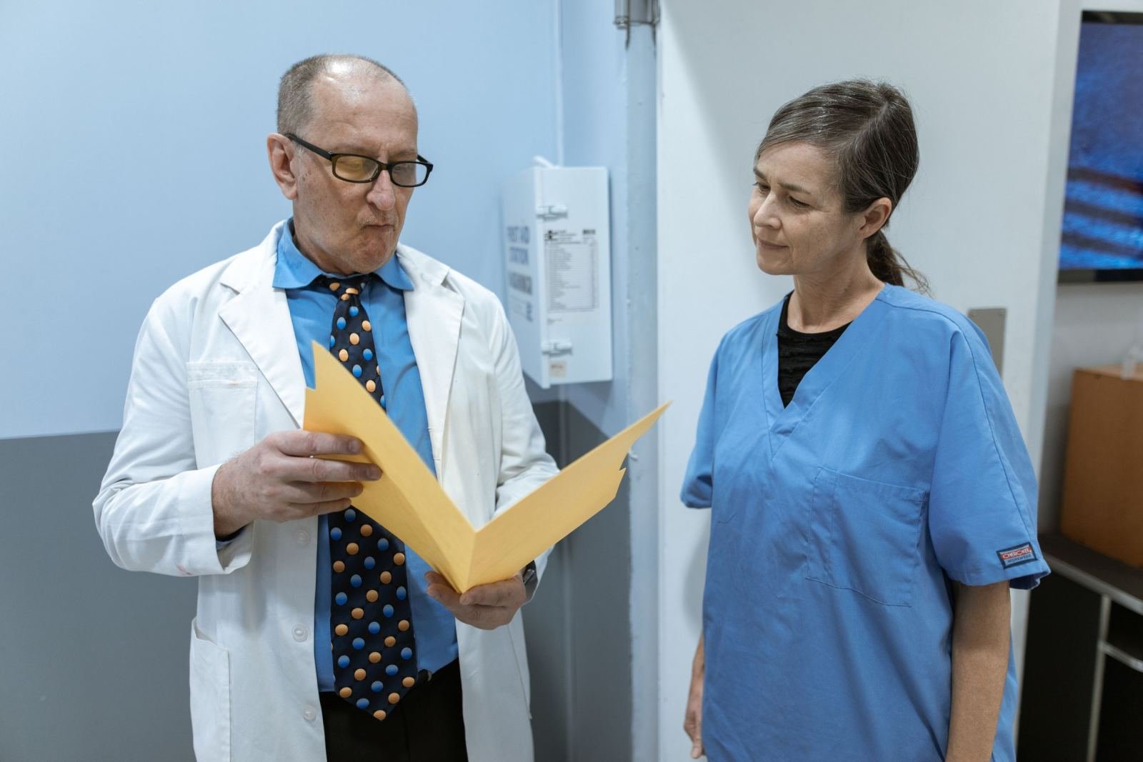 A nurse in blue scrubs and a doctor in a white coat look over a yellow folder.