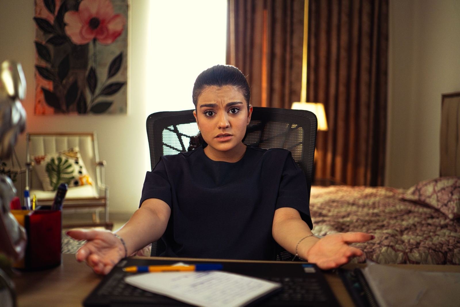 A woman sits at a desk looking confused with her hands open