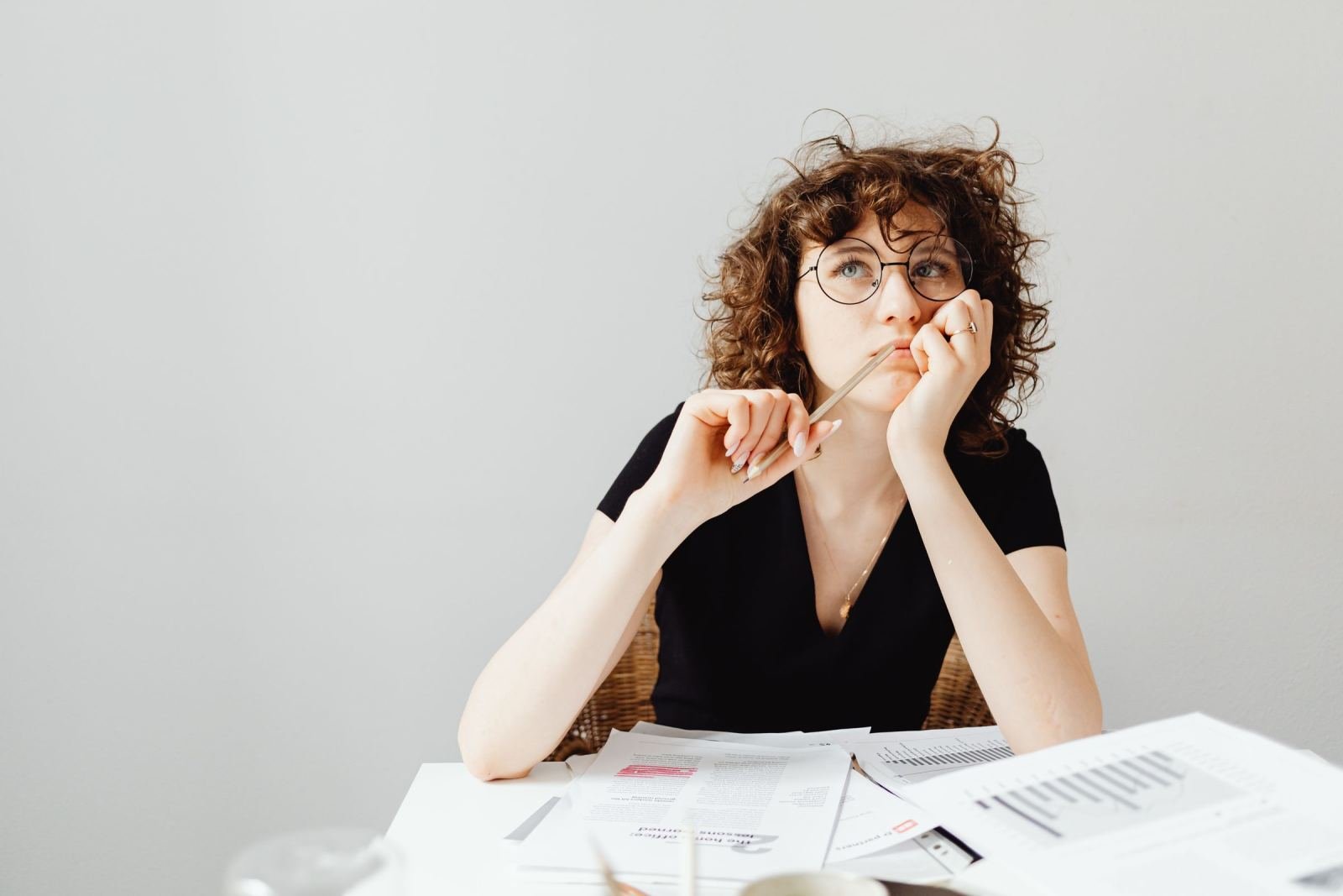 A woman with glasses looks thoughtful while sitting at a table with papers