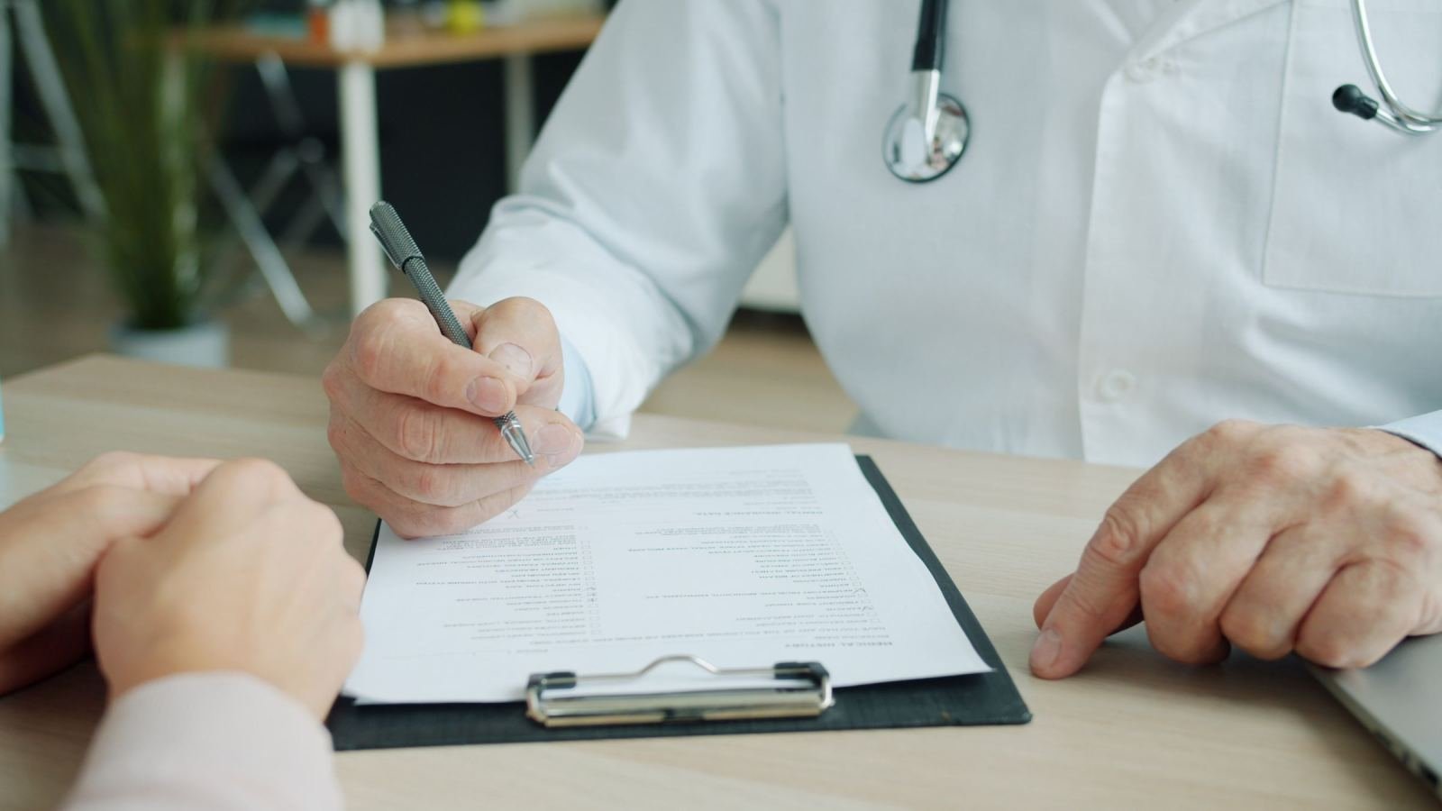 A clinician in a white coat looks at a paper held on a clipboard while talking to a patient.