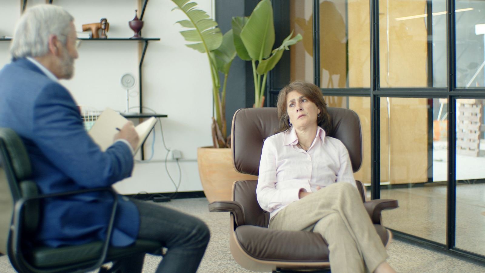 A woman sits in a leather chair while a man with a notebook speaks to her.