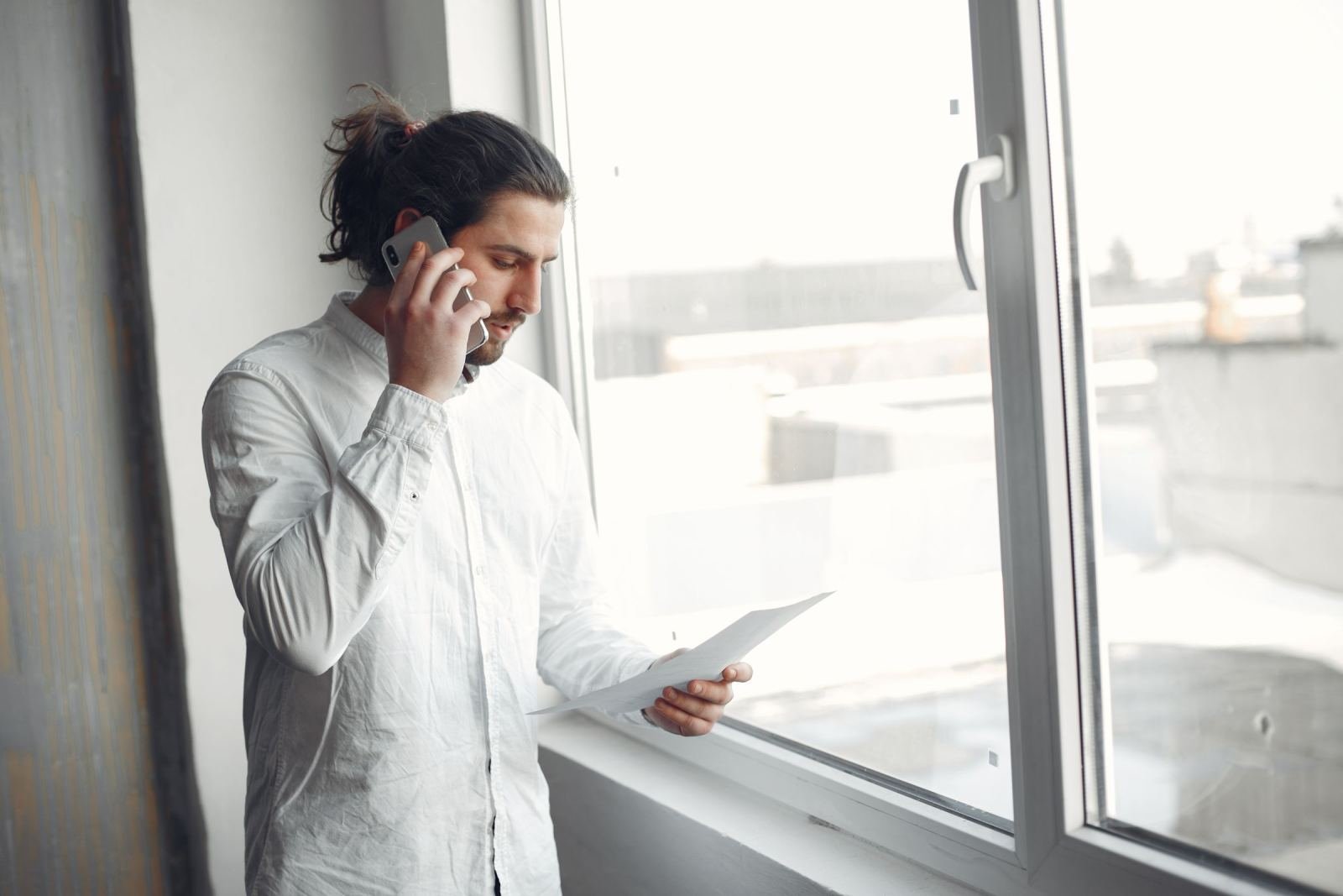 A man talks on a phone while reading a document by a window