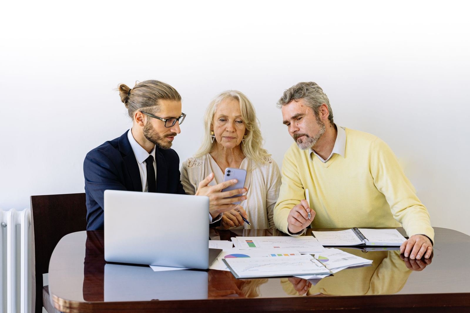 Three people review a smartphone, laptop, and printed charts at a table.