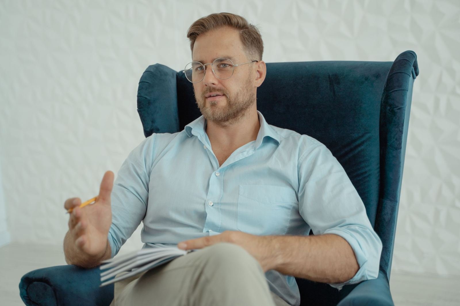 A man with glasses and a notebook sits in a blue velvet armchair