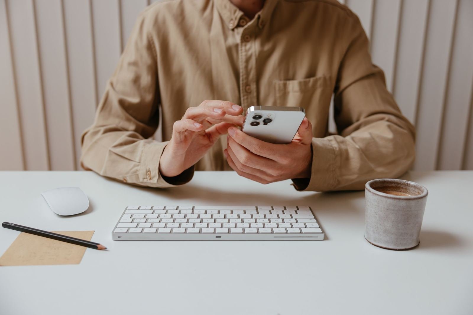 A person in a tan shirt uses a smartphone at a white desk with a keyboard and coffee