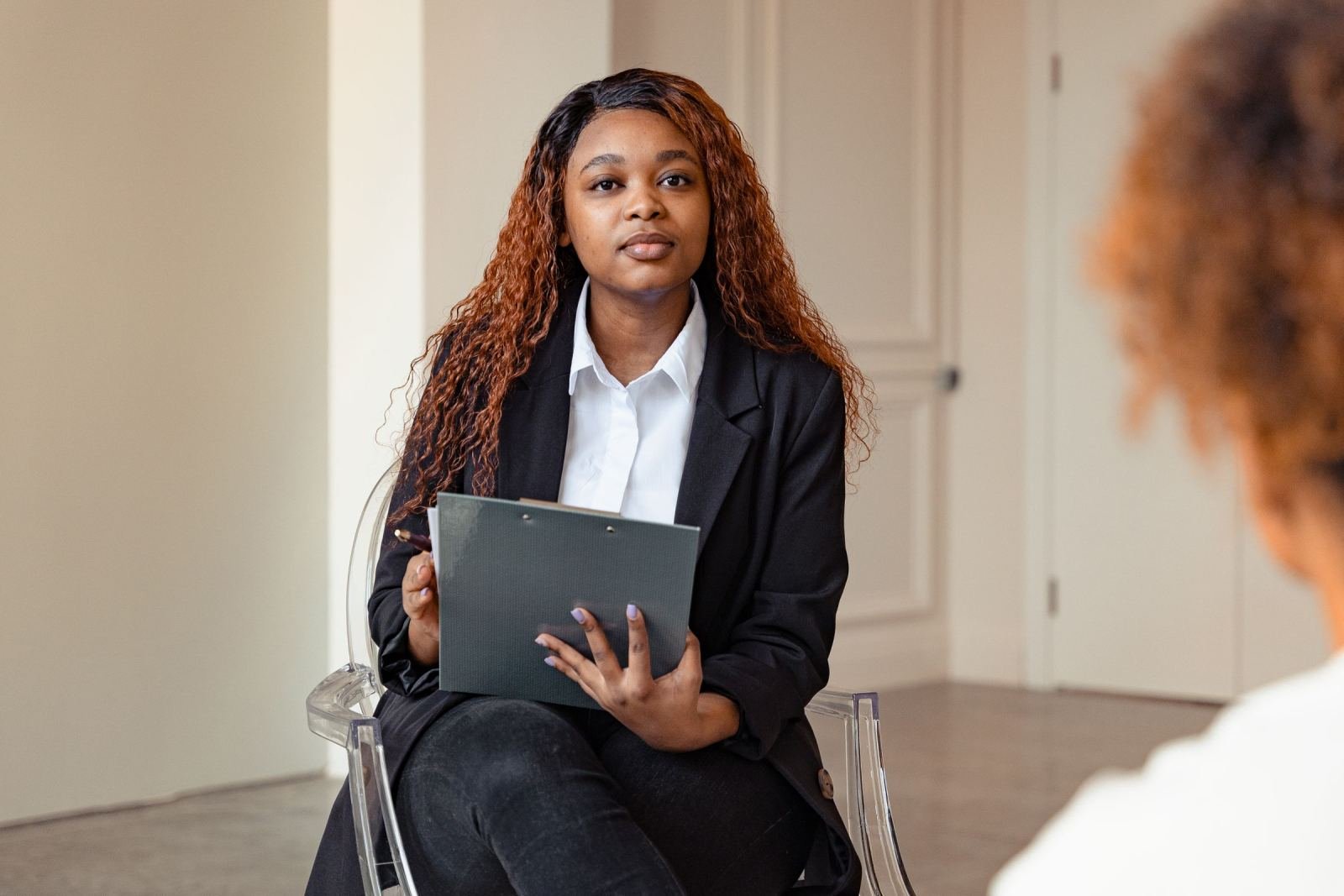 A professional woman holds a clipboard while seated in a transparent chair at a meeting.