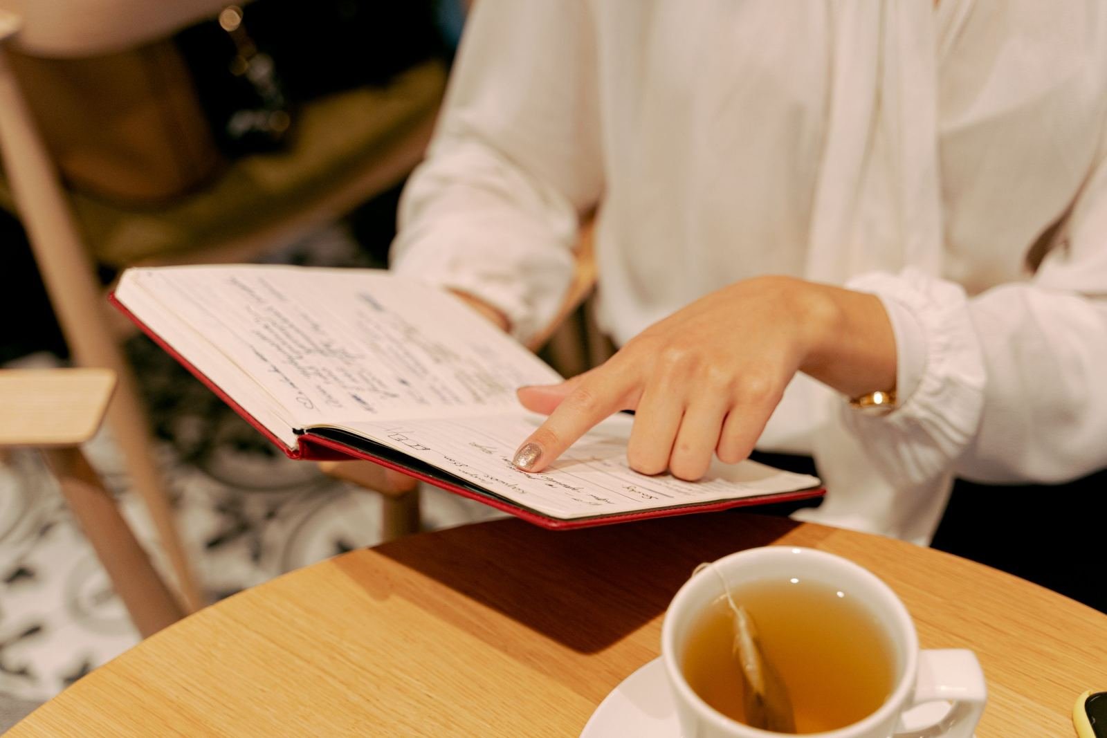 A person in a white blouse points to notes in a red notebook next to a cup of tea