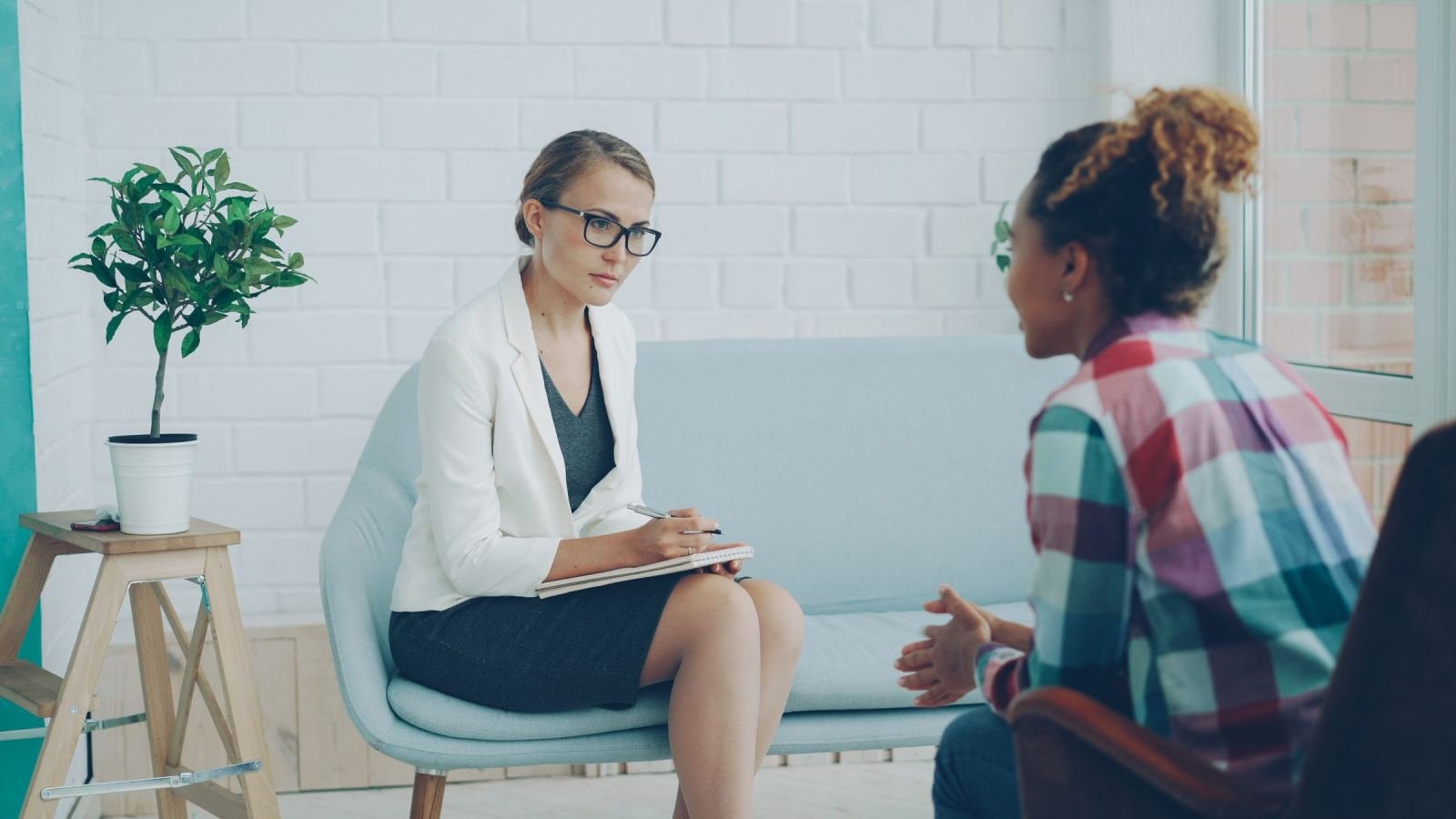 A woman wearing a plaid shirt sits while a counselor takes notes in a notebook.