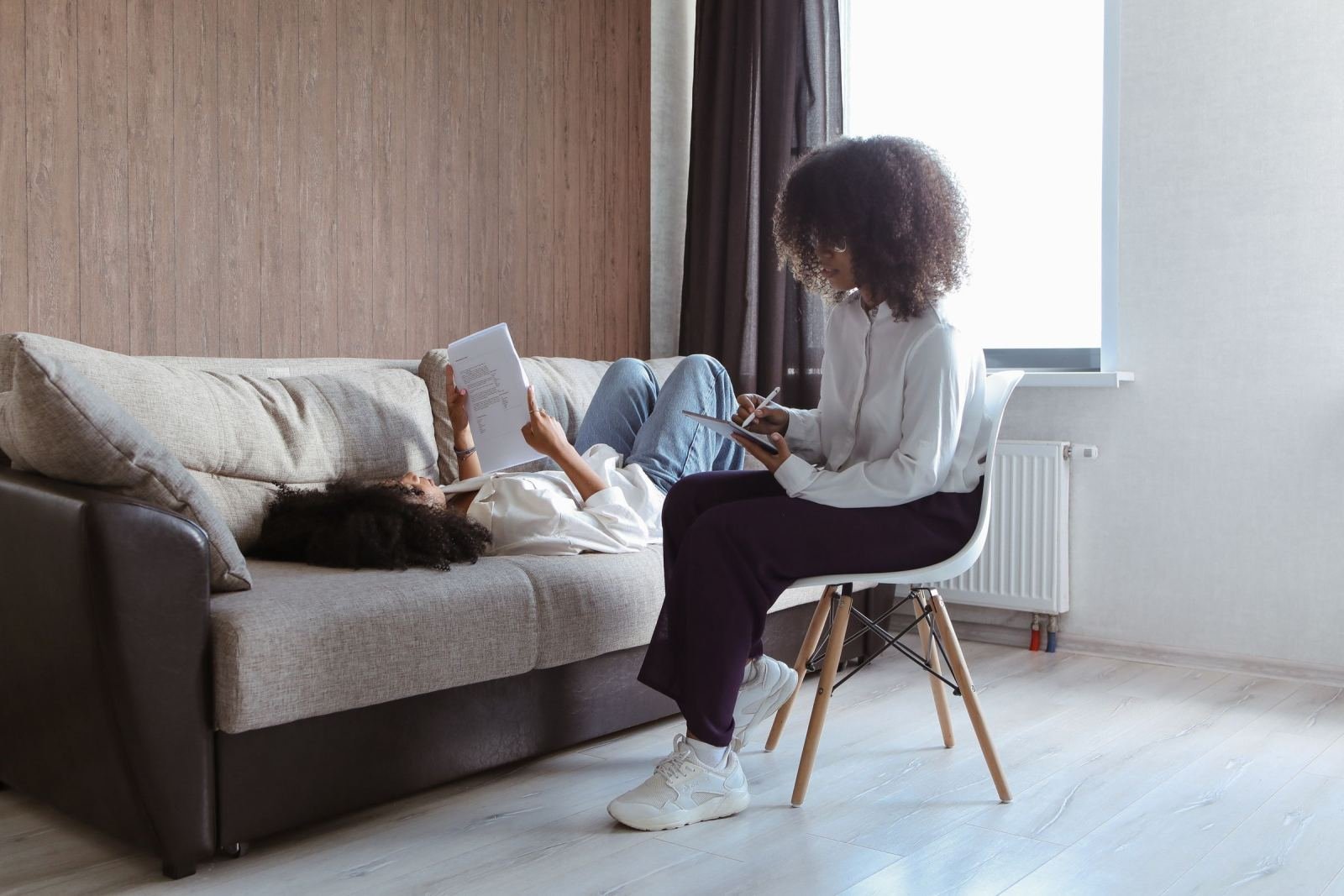 A woman sits in a chair with a notebook while another woman lies on a couch holding a paper