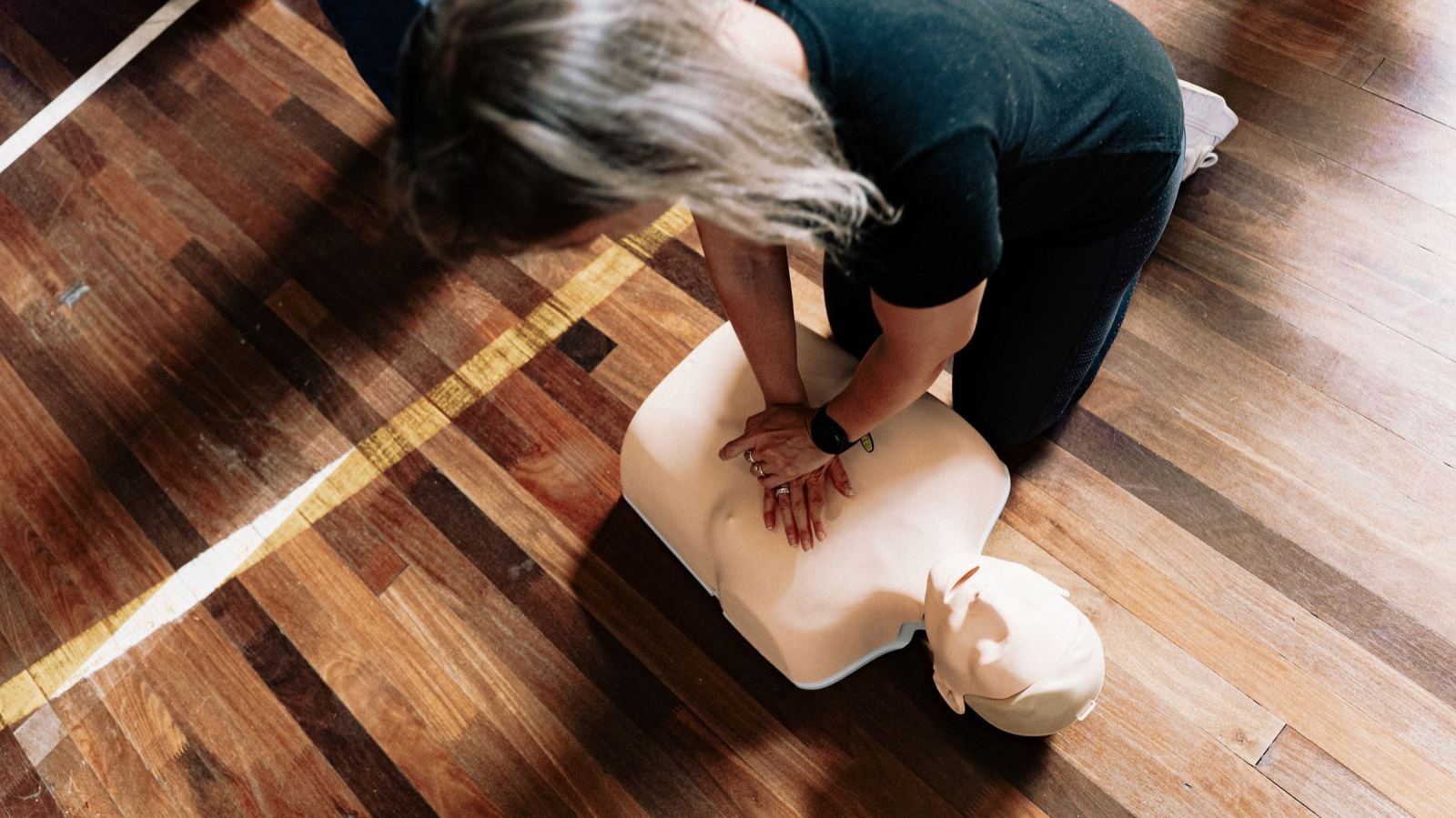 Someone performs chest compressions on a CPR dummy on a wood floor