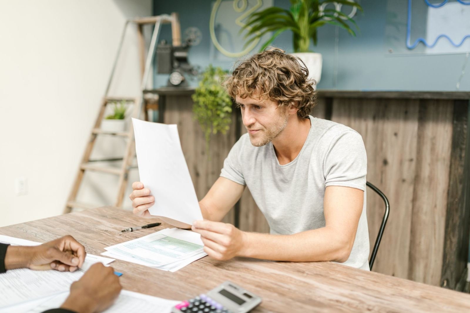 A man in a grey t-shirt reviews a document while sitting at a wooden table with another person