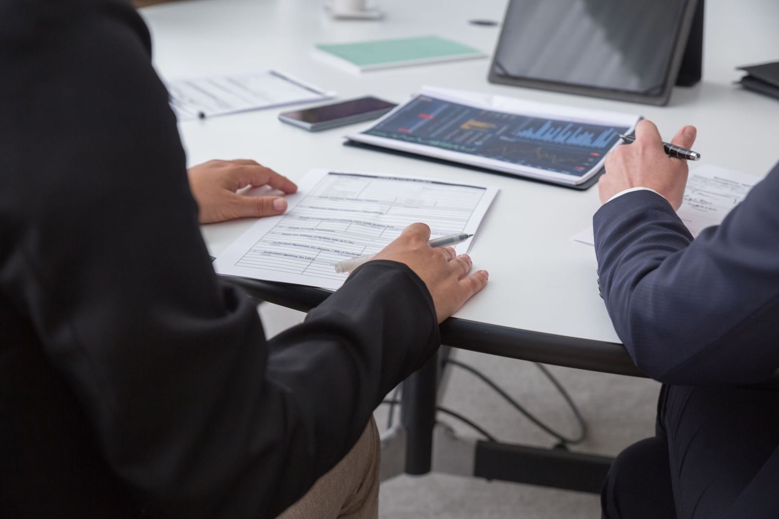 Two people in business suits review documents and a tablet at a white desk