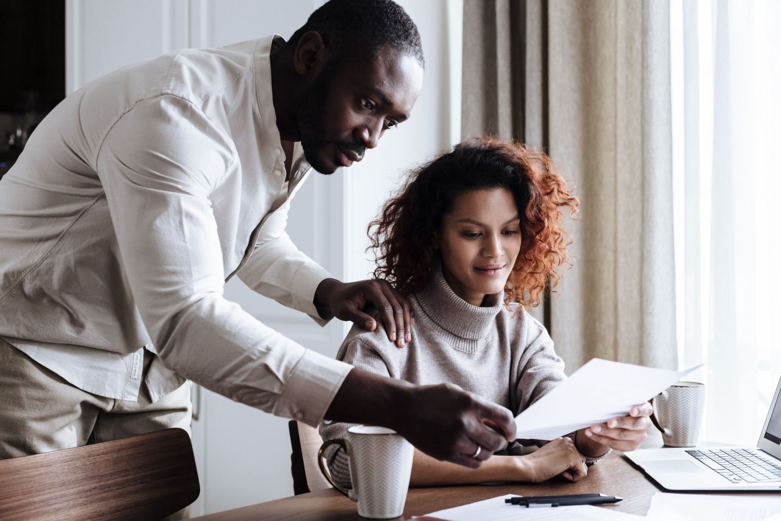 A couple sits at a table with a laptop, looking over a paper together.