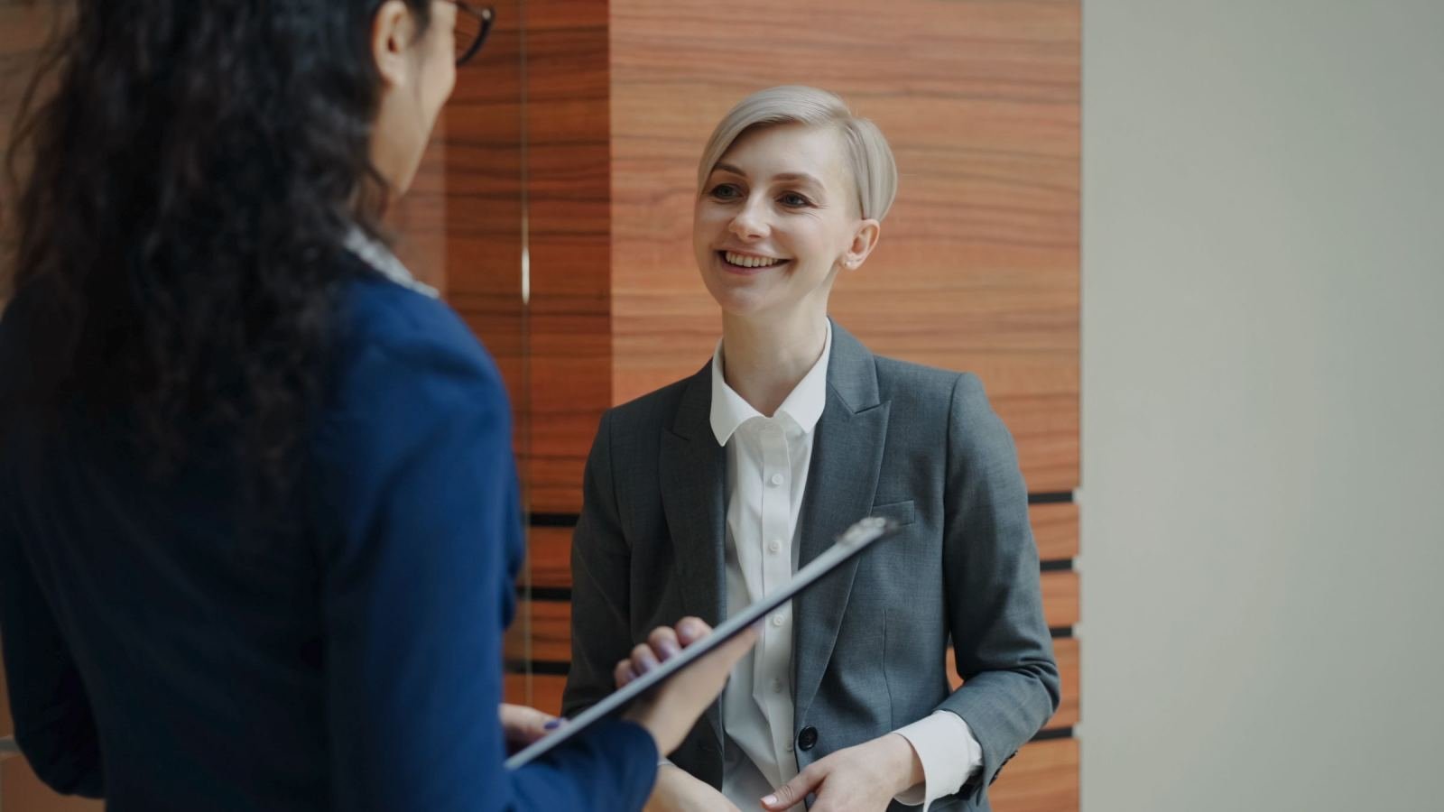Two women in professional attire talk in an office, one holding a clipboard