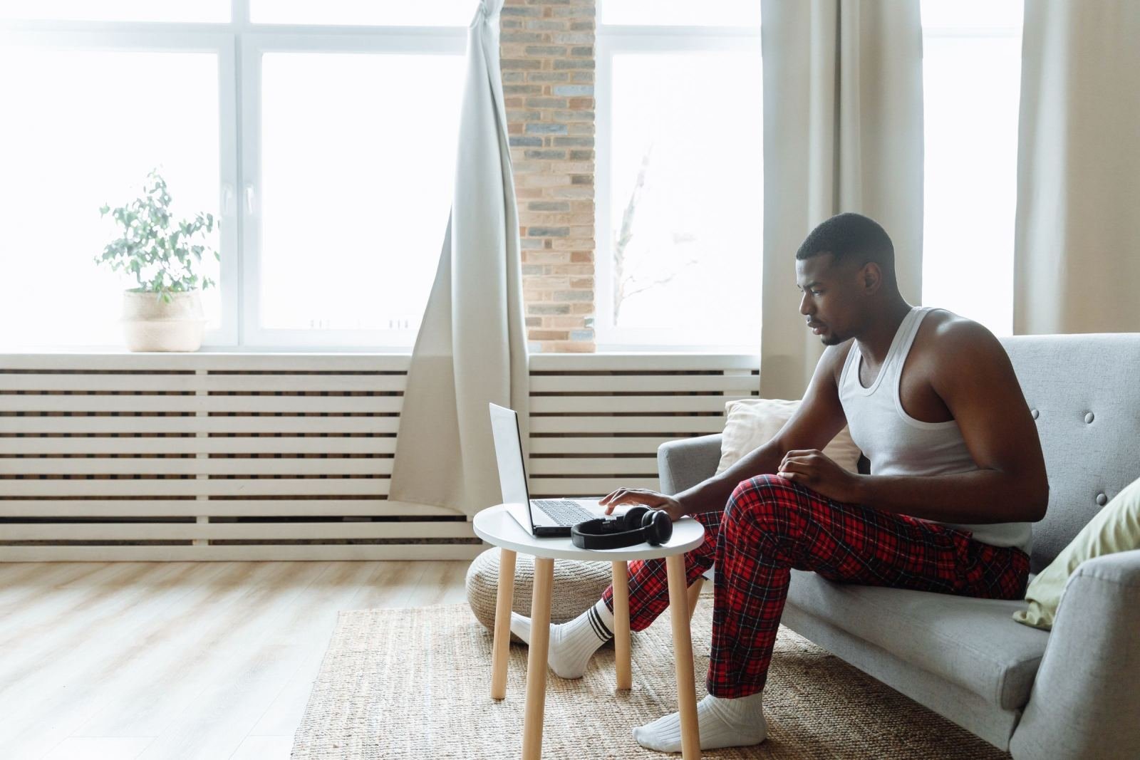 A man sits on a sofa in pajama pants. He works on a laptop on a small table.