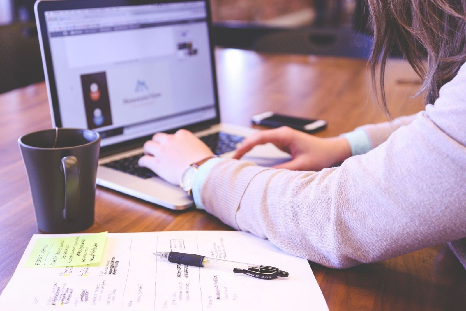 Someone sits at a desk with a laptop, a notebook, and a mug.