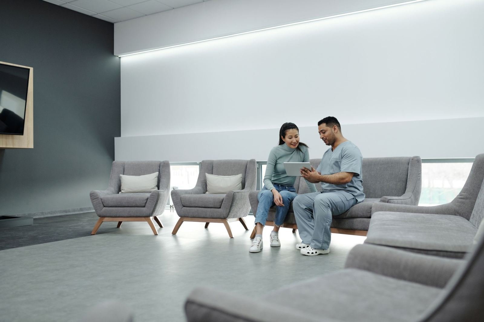 A medical professional in scrubs shows a tablet to a woman in a waiting room