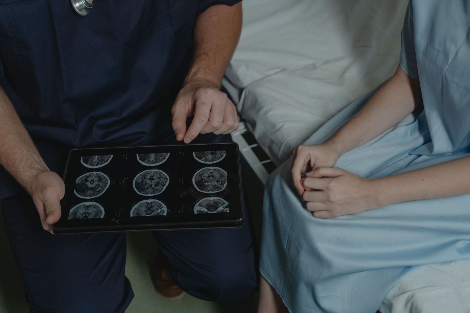 A doctor shows a brain scan on a tablet to a patient wearing a hospital gown.