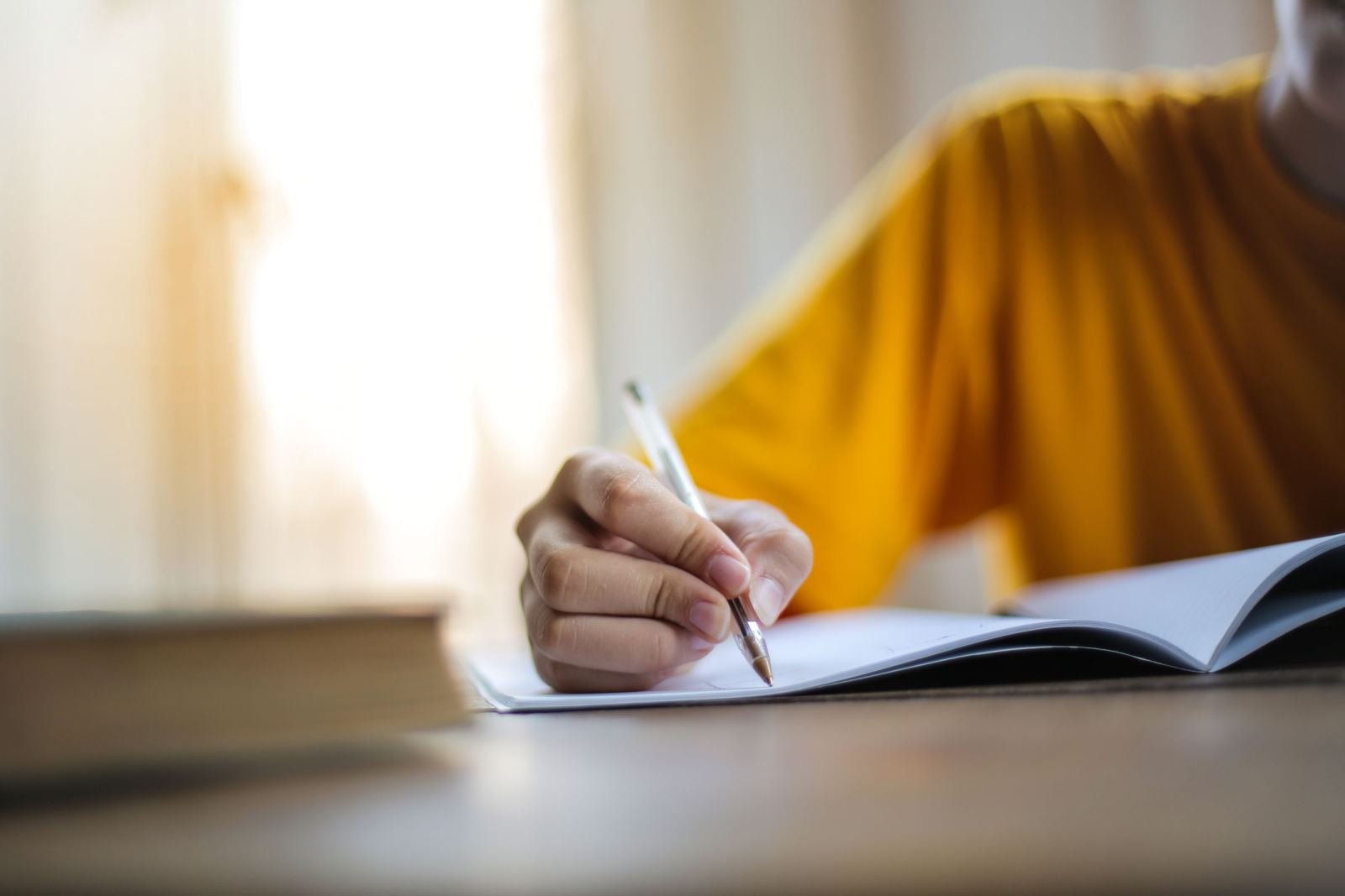 Someone in a yellow top writes in a book on a wood desk.