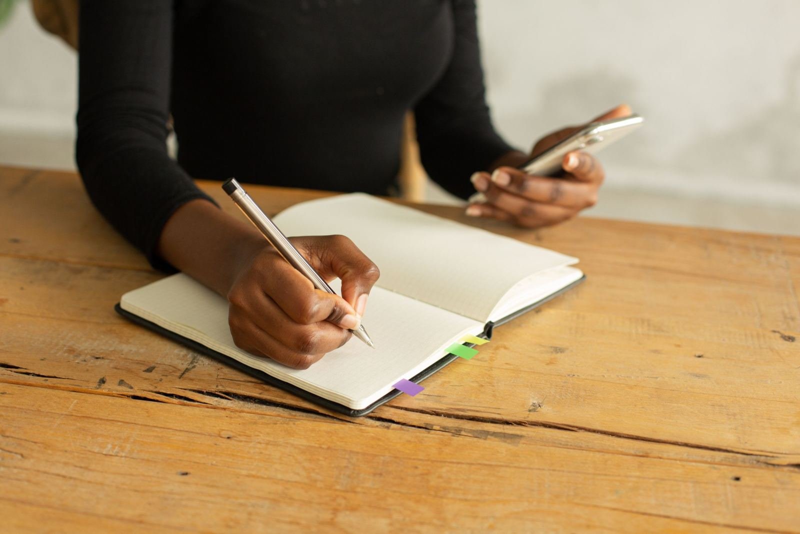 A person writing in a notebook while holding a smartphone at a wooden table