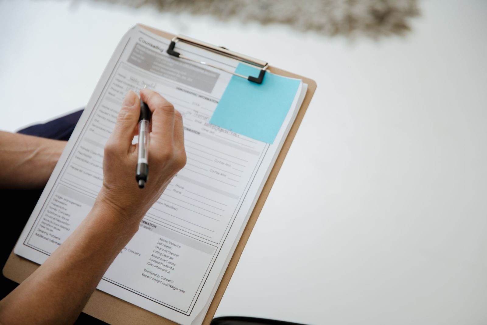 A hand holding a pen over a form on a clipboard with a blue sticky note