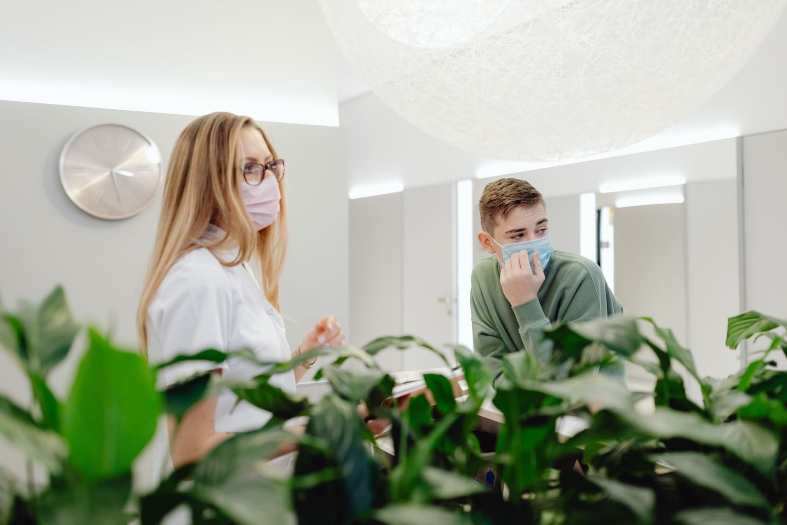 A healthcare provider in a white coat and mask speaks with a patient in a green sweater