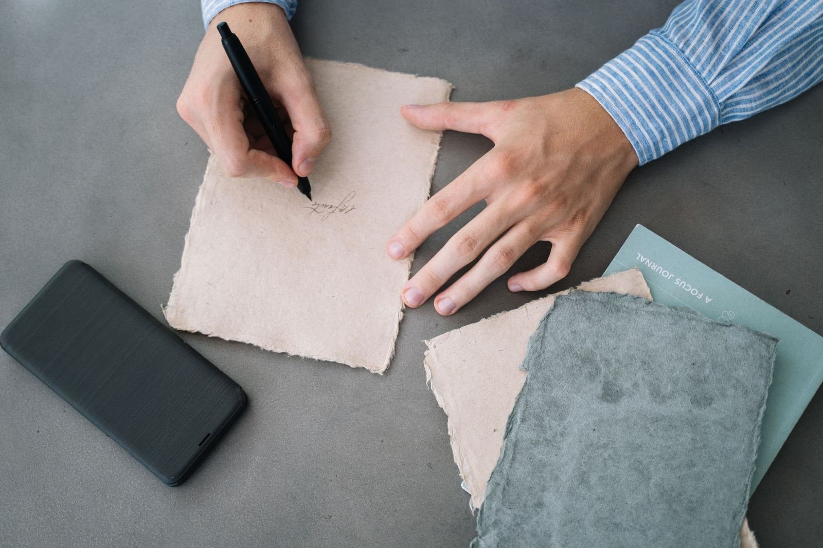 A person writes on handmade paper near a smartphone and a focus journal