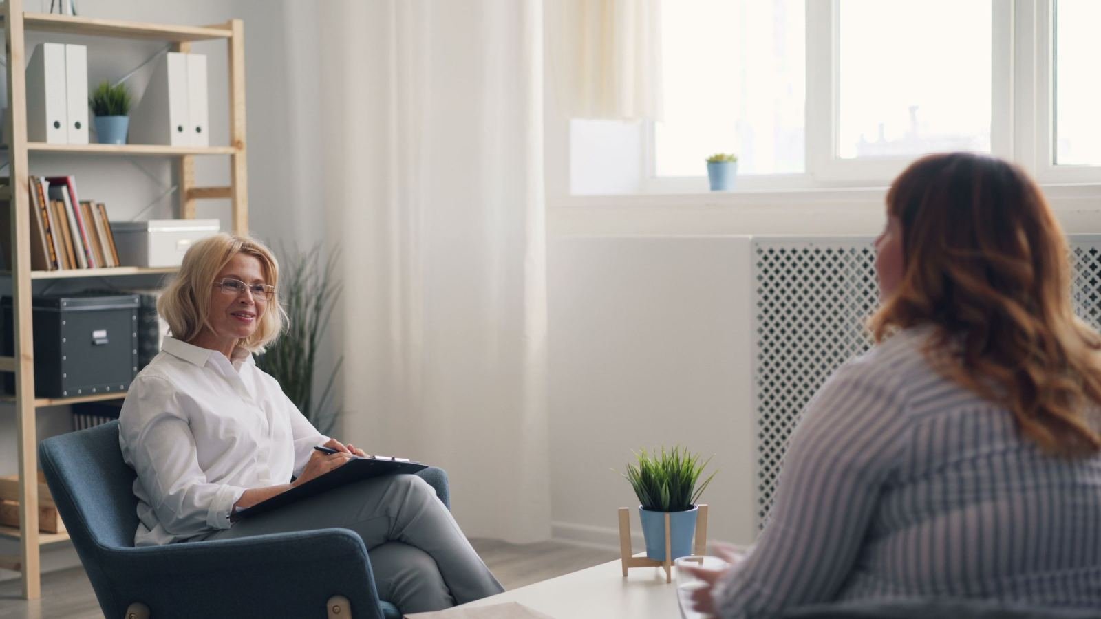 A therapist with a clipboard talks with a client in a bright office