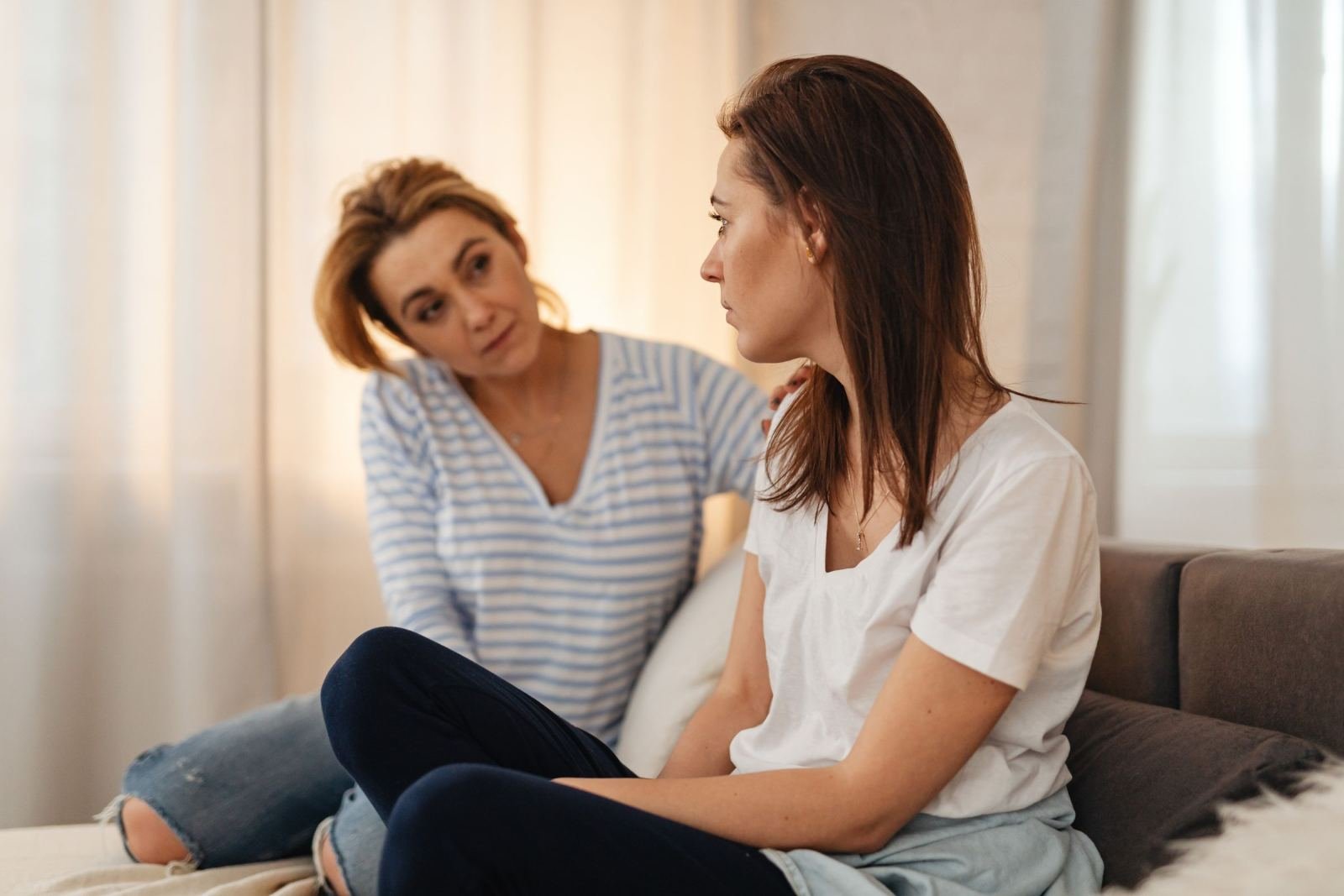 One woman sits on a bed with her hand on the other's shoulder as they talk.