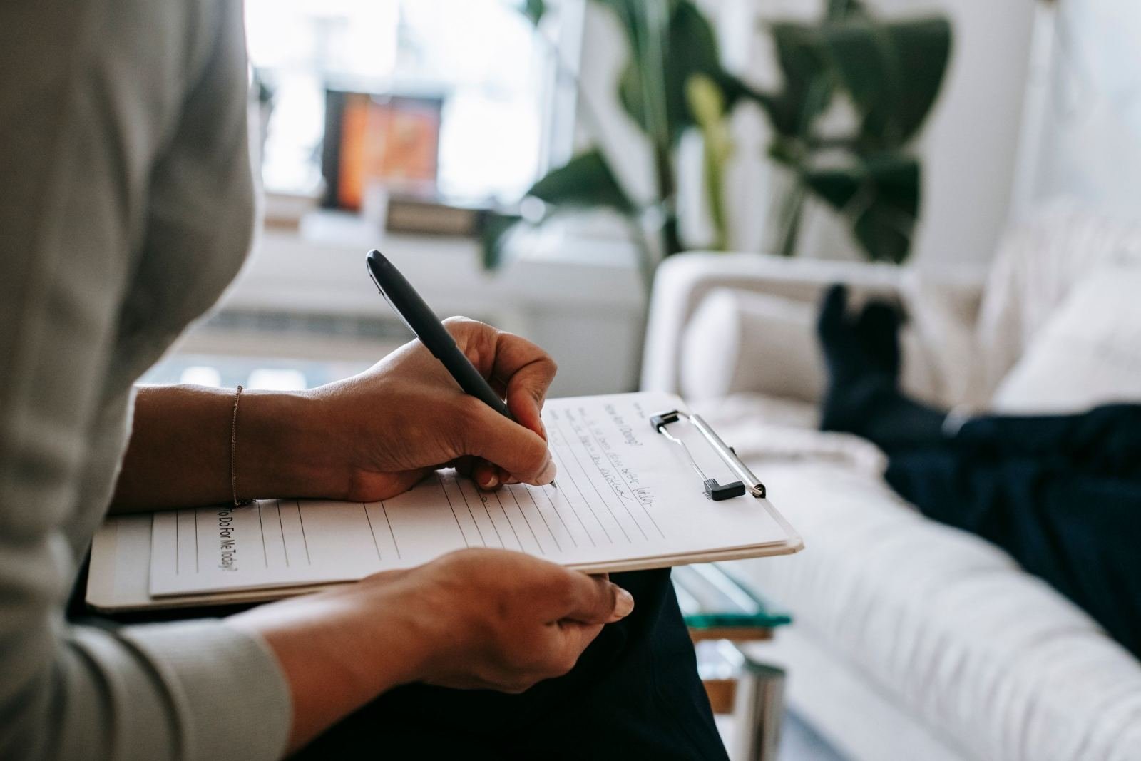 One person sits on a couch while another writes notes on a clipboard.