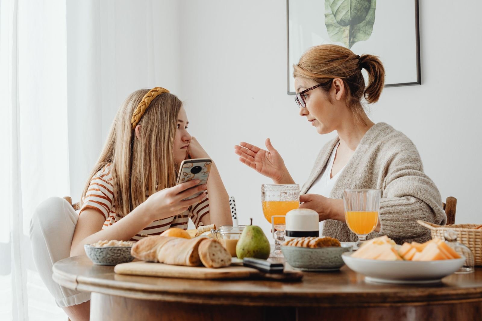 Two women sit at a kitchen table with breakfast and orange juice.
