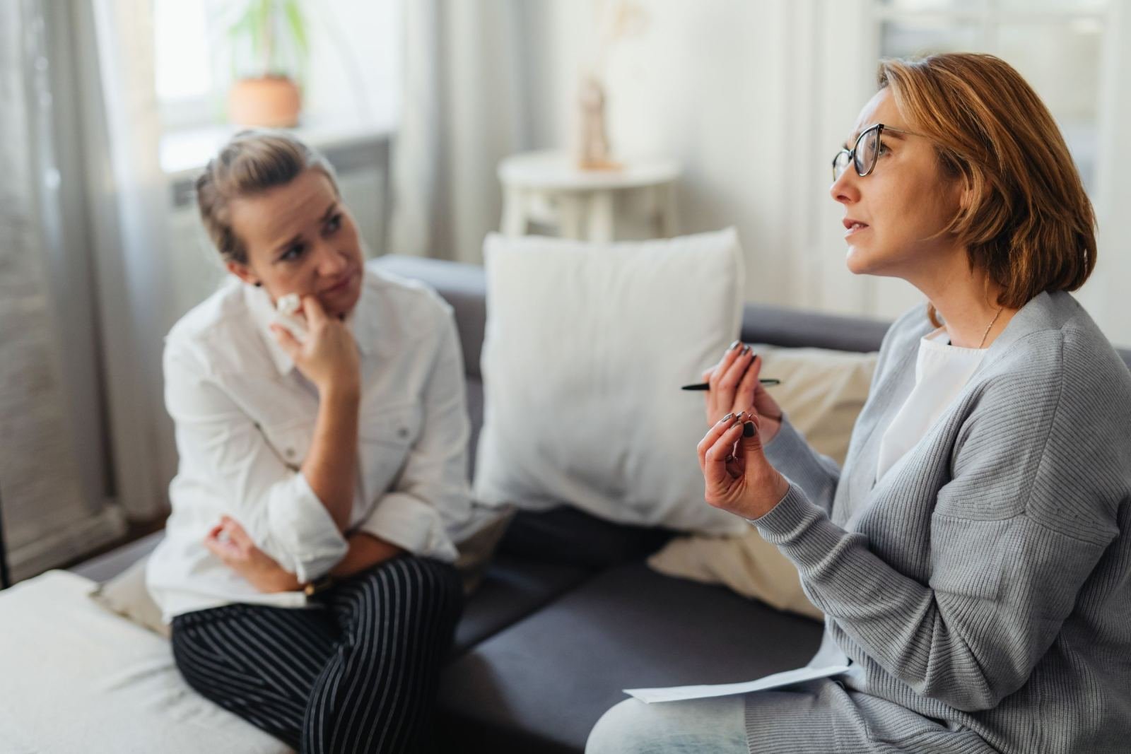 Two women sit on a sofa talking, one gesturing with a pen while the other listens