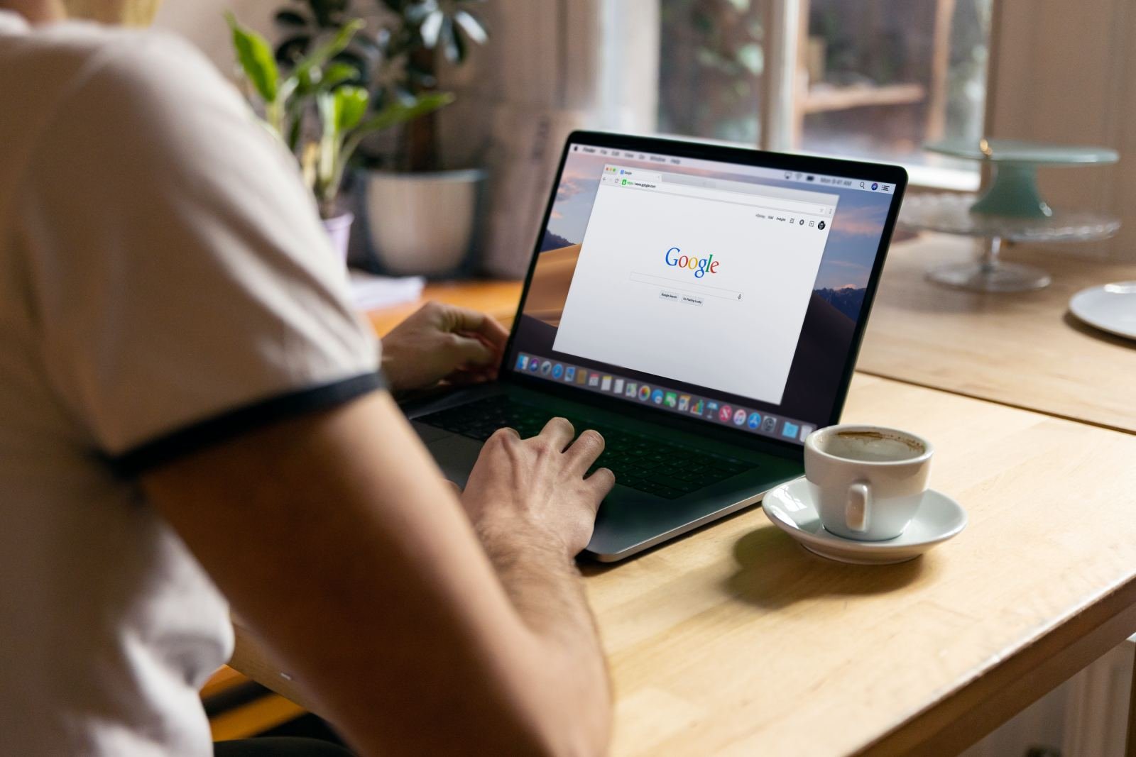 A person sits at a wooden table with coffee. They use a laptop to search the internet.
