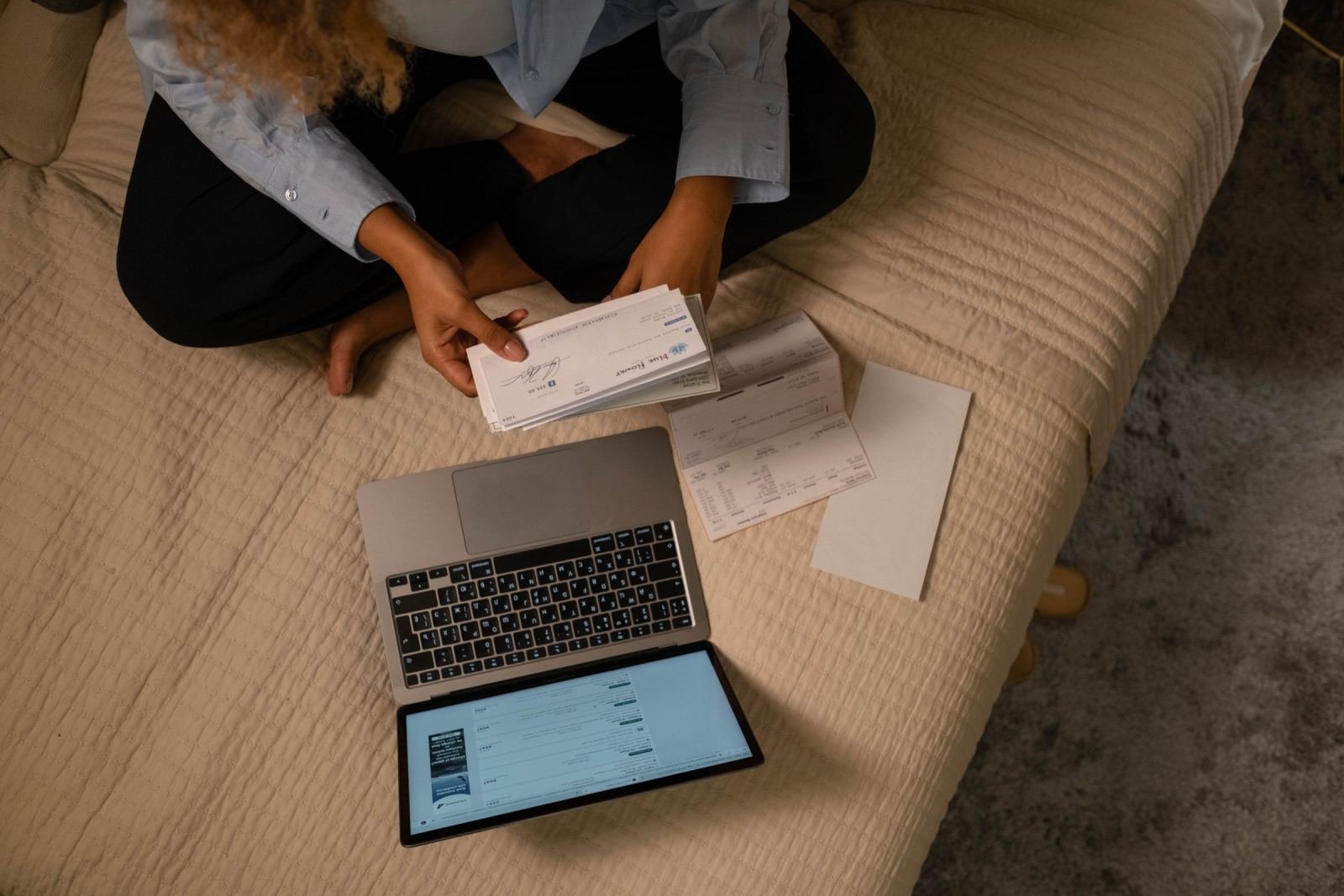 A woman sits on a bed with a laptop open. She is going over paperwork.
