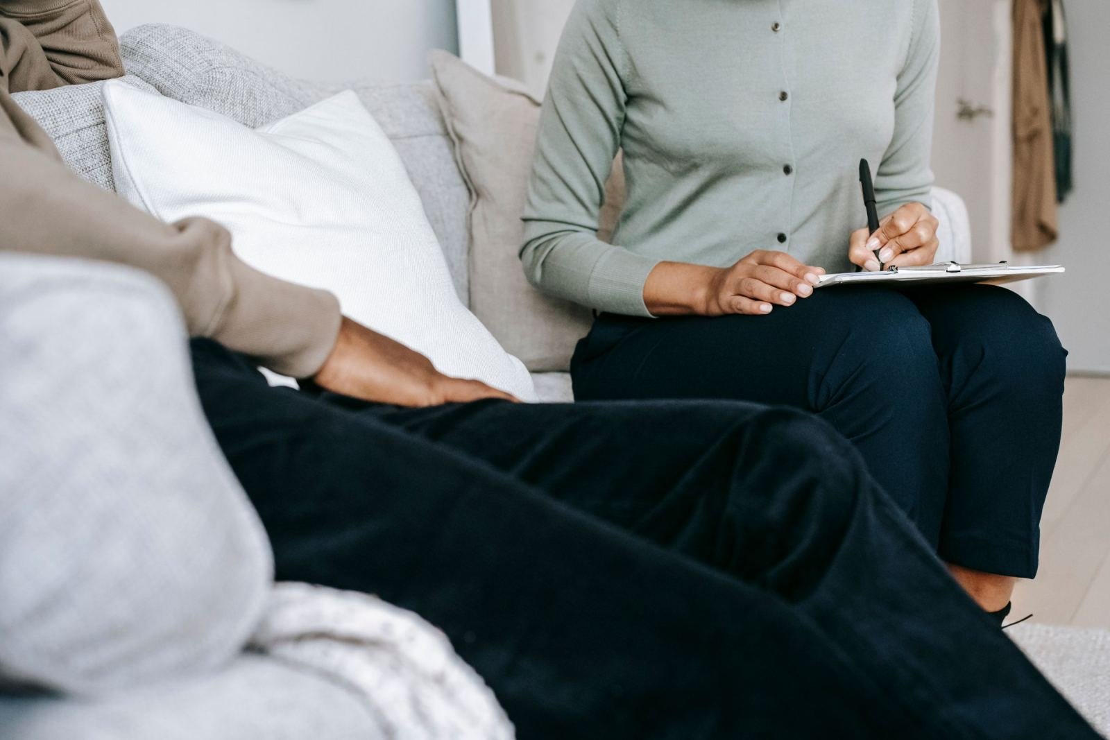 One person sits on a couch taking notes on a clipboard while another sits with them wearing a green shirt.