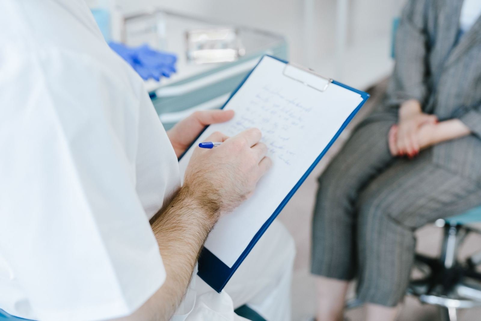 A patient sits close to a clinician in a white coat who marks notes on a clipboard.