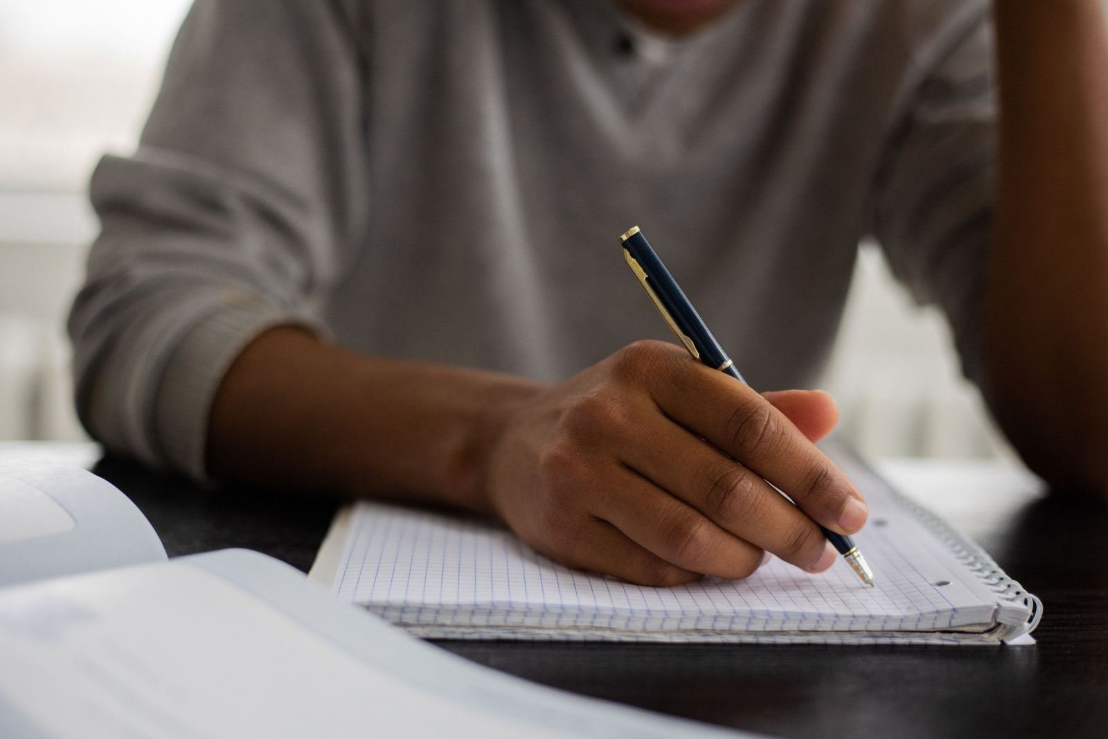 Someone writes in a ruled notebook using a blue pen while sitting at a desk.