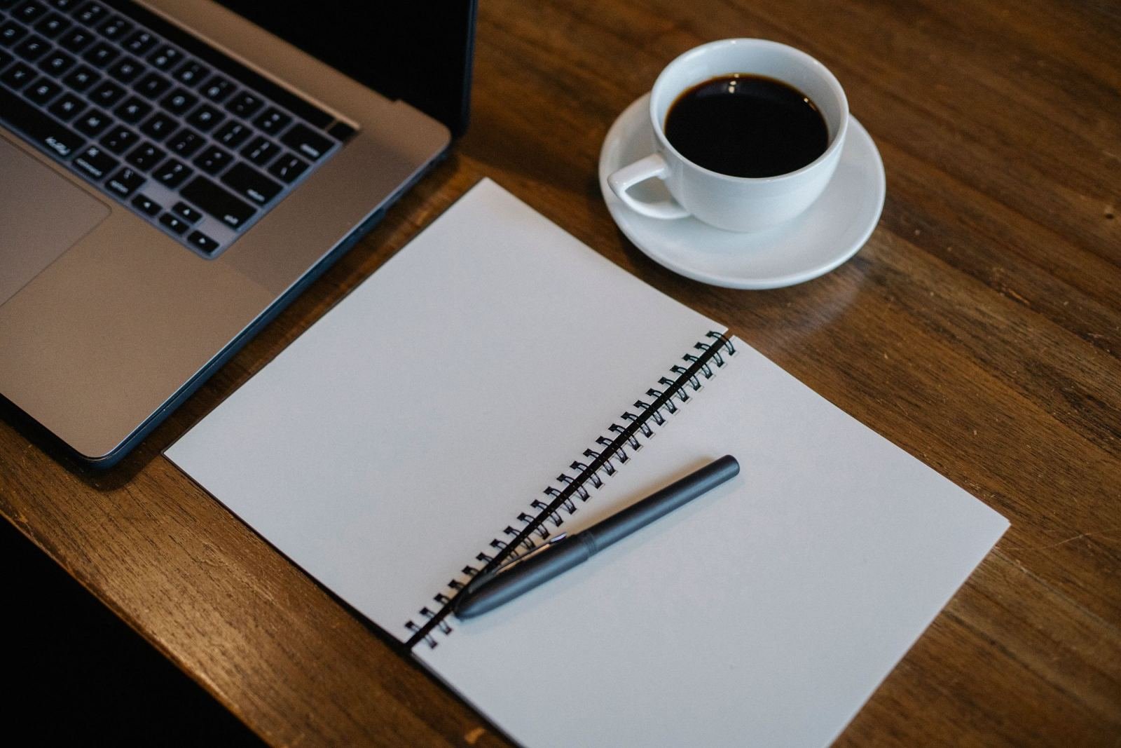 A laptop, a blank spiral notebook with a pen, and a cup of coffee on a wooden desk