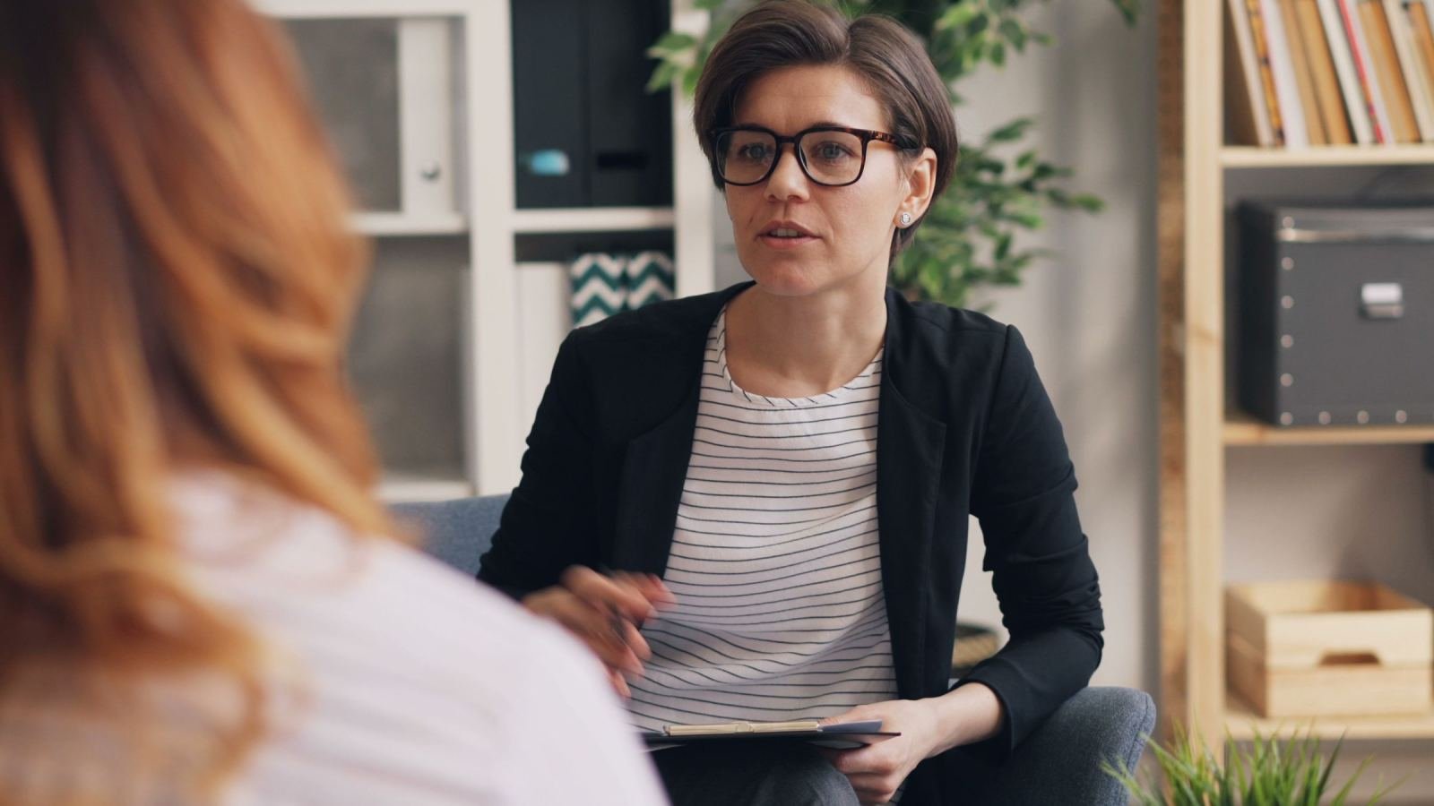 A clinician with glasses speaks to a client in a bright office